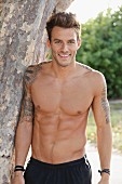 A young, topless, tattooed man standing by a tree