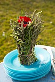 Posy of herbs with red anemone in centre on stack of pale blue plates outdoors