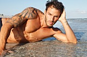 A young, topless, tattooed man lying on a sandy beach