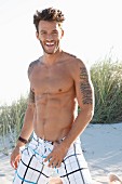A young, tattooed man on a beach wearing bathing shorts