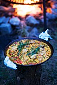 A pan of paella on a tree stump with a fire pit in the background