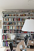 Desk below pendant lamp with white lampshade and metal bookshelves in background