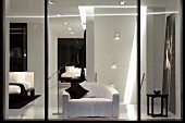 View through glass wall of armchair with white loose cover and black scatter cushions in front of staircase underside with recessed spotlights