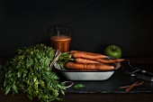 A bunch of carrots in an enamel bowl with carrot juice and an apple