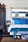 Blau-weiss gemusterte Kissen auf Bett vor weisser Holzwand mit Vintage Spiegel, an Bettende blaue Kleiderbank