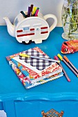 Colourful exercise books and felt-tip pens in front of retro teapot used as pen holder on blue table