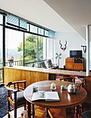 Round, wooden, antique table and armchairs in front of free-standing, wooden sideboard; hunting trophies on wall of lounge area in background