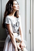 A young woman standing in front of a cupboard holding a teddy bear and wearing a T-shirt and a skirt