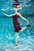 A woman wearing a red dress and high heels standing on one leg under the water in the swimming pool