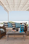 Coffee table in front of sofa with striped scatter cushions below pergola with fabric canopy; beach and sea in background