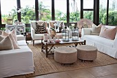Comfortable lounge area in conservatory: sofas with white loose covers, arranged scatter cushions and pouffes around coffee table
