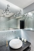 Kugelförmiges Waschbecken auf Granitplatte vor Wand mit Mosaikfliesen, oberhalb Designerleuchten