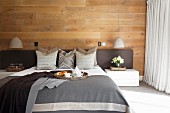 Double bed with grey covers, upholstered headboard, scatter cushions and throws in bedroom with wood-clad wall