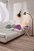 Modern double bed with white headboard and round, colourful scatter cushions; folding bedside table and retro standard lamp