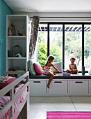 Mädchen beim Spielen, auf eingebauter Bank mit Polster vor Fenster in modernem Kinderzimmer