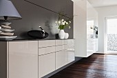 Weisses Designer-Sideboard, im Vordergrund Tischleuchte vor grauer Wand, im Hintergrund Kücheneinbauschrank mit Geräten