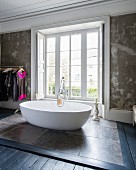 Oval, free-standing designer bathtub on stone tiles in front of bedroom window