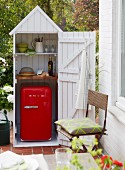A white-painted mini shed as a kitchen cupboard for a terrace for crockery and a retro fridge