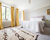 View across rug to double bed and yellow floor-length curtains on windows in simple bedroom