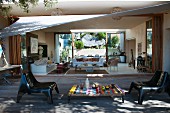 Terrace with awning, low colourful table, modern plastic chairs and view into interior of house