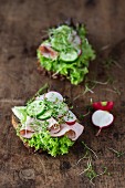 Two open sandwiches with lettuce, ham, cucumber and radishes on a wooden surface