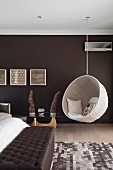 White, wicker, hanging chair in bedroom with wall painted dark brown