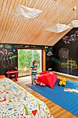 Kleiner Junge neben rotem Sitzsack auf blauem Teppich, an Wand Tafellack und Kreidezeichnungen, im Kinderzimmer unter dem Dach, holzverkleidete Decke