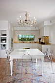 Ghost chairs around white, country-house-style dining table below chandelier in open-plan kitchen