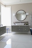 Elegant bathroom with glossy floor, washstand with grey drawers and large round mirror