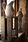 Old washstand pitcher on wooden crate in front of religious statue on windowsill