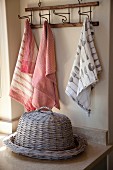 Flat, wicker basket with wicker cover on worksurface in front of tea towels hanging from hooks