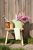 Pastellgrüne Giesskanne mit Fliederstrauss und Aufbewahrungsbehälter auf Holzschemel, im Freien vor Holzwand