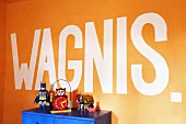 Botschaft auf orangefarbener Wand, davor teilweise sichtbarer blauer Schrank mit Vintage Spielsachen