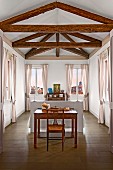 Antique desk and chair behind bed headboard, windows with pale pink curtains and exposed, rustic roof beam structure