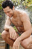 Mann in Badeshorts hockt unter Dusche im Freien