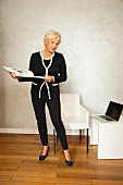 An older business woman with a coffee-table book standing next to a laptop
