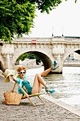 Junge Frau im Liegestuhl an Fluss vor Steinbrücke