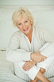 An older woman wearing white pyjamas and a cardigan sitting on a bed