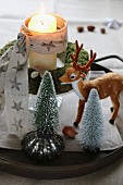 Dekoidee zu Weihnachten, Miniatur-Tannenbäumchen und Rentierfigur vor Adventskranz mit brennender Kerze im Glas