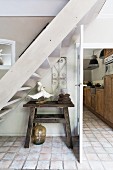 Rustic wooden table below white-painted wooden staircase in hallway with pale terracotta floor and view into kitchen through open door to one side