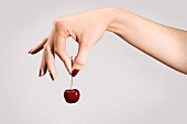 A woman's hand holding a cherry by the stem between her thumb and forefinger