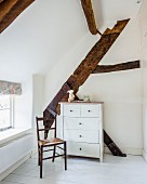Black-painted wooden chair and white chest of drawers in front of exposed wooden roof beams in attic room