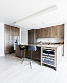 Black bar stools at free-standing counter with integrated wine cooler in designer kitchen with wooden cupboard doors
