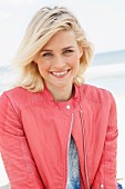 A blonde woman by the sea wearing a blue jumper and a salmon coloured leather biker jacket