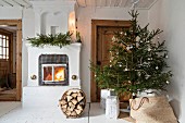 Decorated Christmas tree next to fire in old fireplace and basket of firewood on floor in rustic interior