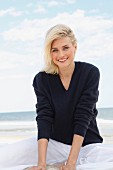 A blonde woman on a beach wearing a dark woollen jumper and white trousers