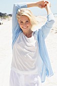 A blonde woman on a beach wearing a white shirts, trousers and a light blue cardigan