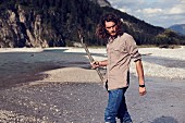 A young man with a beard and long hair walking along a gravelly river bed