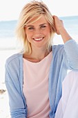 Lächelnde blonde Frau in pastellfarbenem Shirt, Jacke und Hose sitzt am Strand