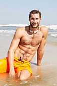 A muscular young man wearing yellow Bermuda shorts sitting in the shallows on a beach